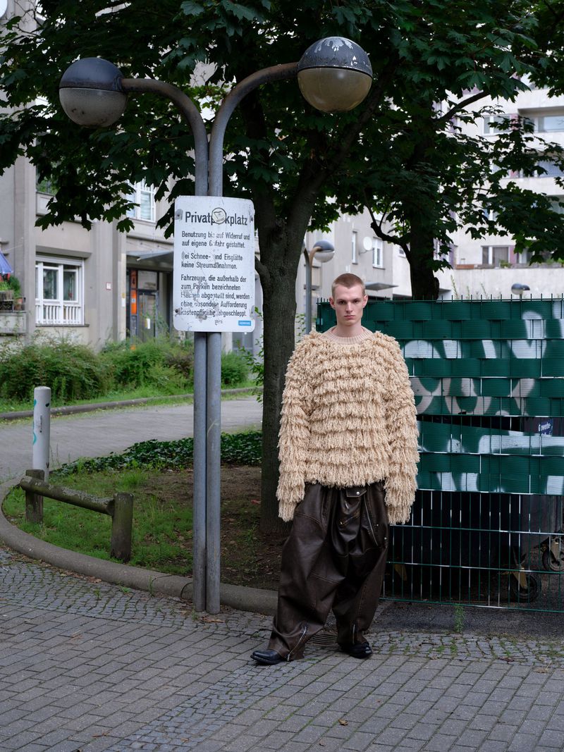 A person with light skin stands outdoors wearing a beige, fringed sweater and shiny brown, oversized pants with a street and greenery.