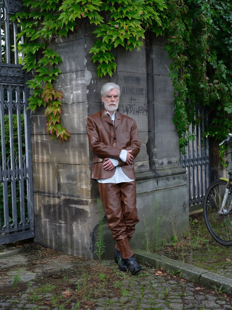 Stylish elderly man in brown leather suit poses against a weathered stone wall with ivy. Outdoor fashion, silver hair, and formal wear.