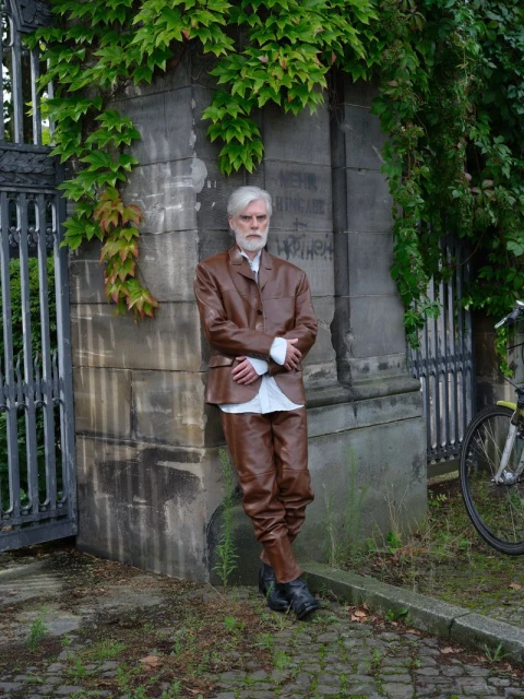 Stylish elderly man in brown leather suit poses against a weathered stone wall with ivy. Outdoor fashion, silver hair, and formal wear.