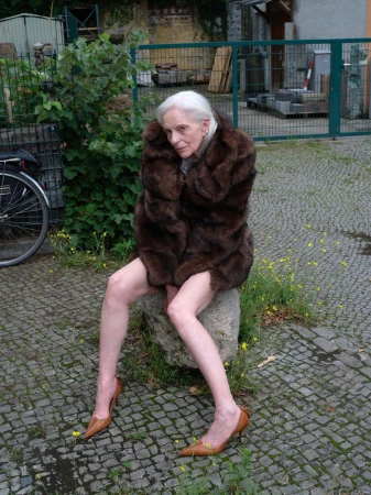 Elderly woman in fur coat poses on stone outdoors. She wears tan heels, showing legs against a leafy and cobblestone backdrop.