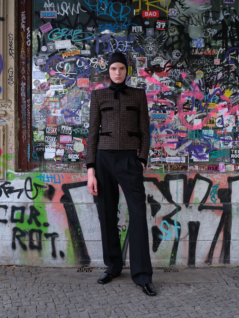 Adult fashion model stands in black beanie, jacket, and pants against a colorful graffiti wall. Outdoor fashion portrait.