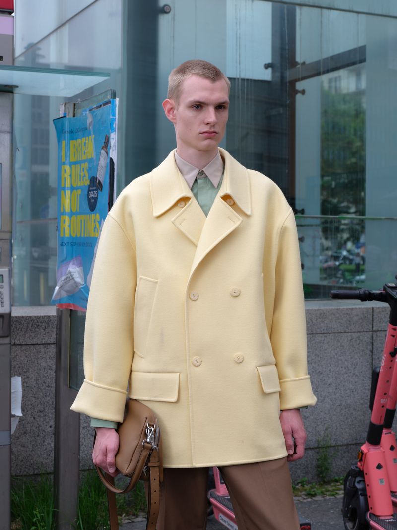 Man in yellow coat poses outdoors in urban setting with scooter. He carries a bag with a strap over his shoulder. 