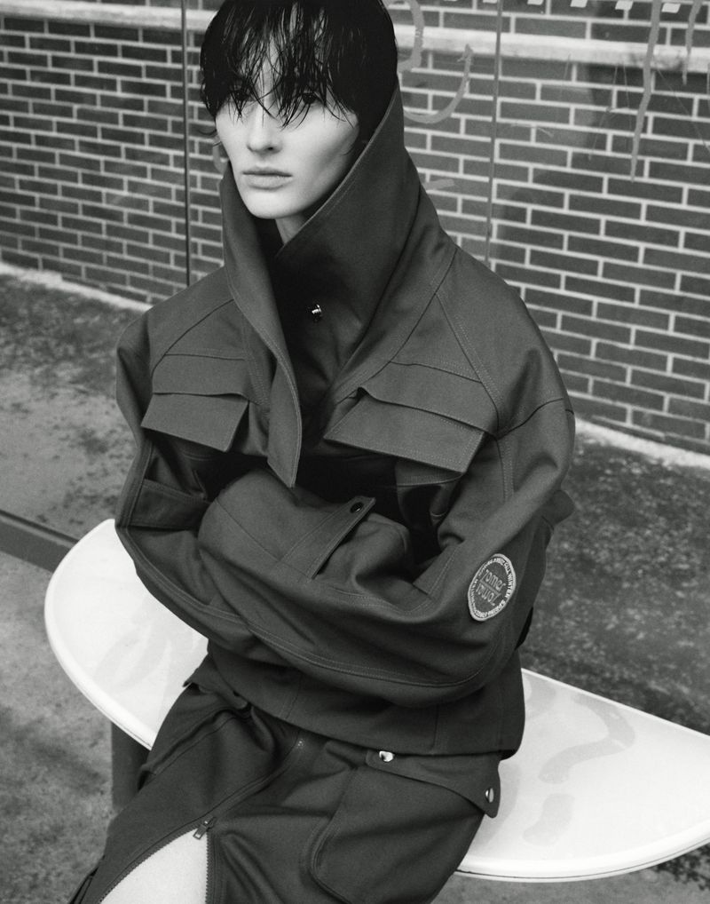 Fashion shot of a person wearing a dark green, high-collared jacket and matching pants, sitting on a white stool in front of a brick wall.