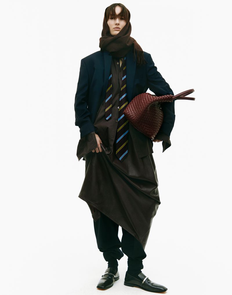 A person in dark clothing poses in a studio. The person is wearing a jacket, skirt, and scarf. They are holding a dark purse.
