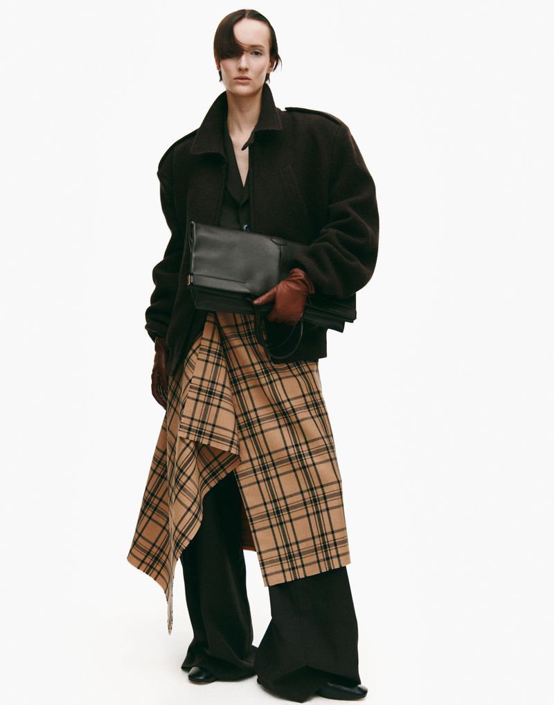 A stylish woman in a dark coat holds a black clutch with a beige plaid skirt, set against a clean white backdrop for a fashion-forward aesthetic.