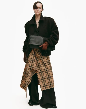 A stylish woman in a dark coat holds a black clutch with a beige plaid skirt, set against a clean white backdrop for a fashion-forward aesthetic.