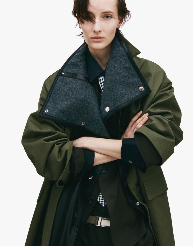 Fashion shot of a woman in a green coat and dark suit, captured in a studio setting with a clean white backdrop and soft lighting.