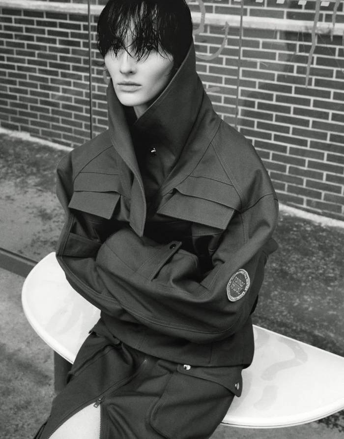 Model with wet dark hair wearing a structured utility jacket seated outdoors against a brick wall.