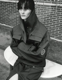 Model with wet dark hair wearing a structured utility jacket seated outdoors against a brick wall.