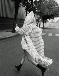 Model Mauve Dupuy completely draped in an oversized patterned hooded cape while running down an urban street in black and white.