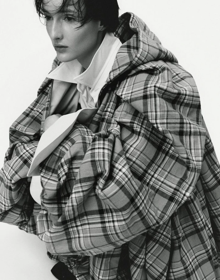 Model wearing a plaid hooded coat over a white shirt against a plain white background for T - Le magazine du Temps
