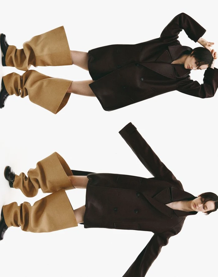 Model Mauve Dupuy in an oversized dark coat and wide camel leg covers against a white seamless studio backdrop.