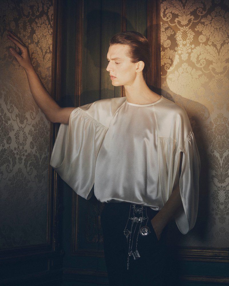 Elegant portrait of a young adult male in a white satin blouse and black pants, posed against an ornate patterned wall indoors.