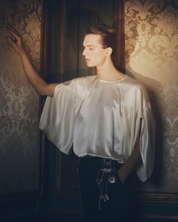 Elegant portrait of a young adult male in a white satin blouse and black pants, posed against an ornate patterned wall indoors.