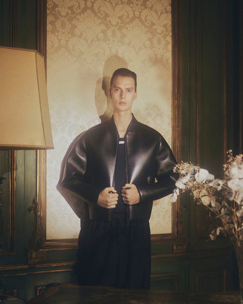 A young adult man poses indoors wearing a dark leather jacket, with a wall lamp and floral wallpaper in the background.