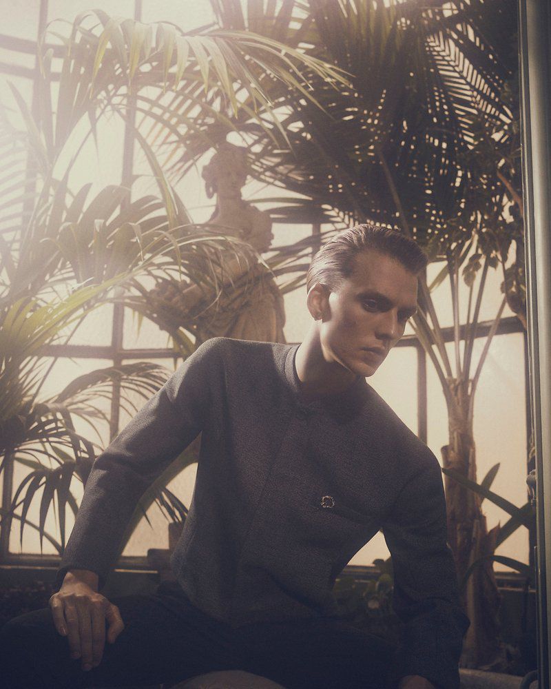 Man in grey shirt poses amid lush indoor greenery, seated against a warm, softly-lit background with a classical statue.