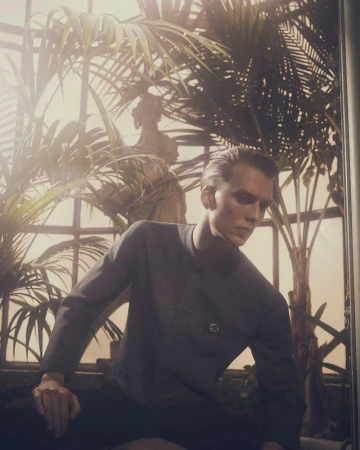 Man in grey shirt poses amid lush indoor greenery, seated against a warm, softly-lit background with a classical statue.