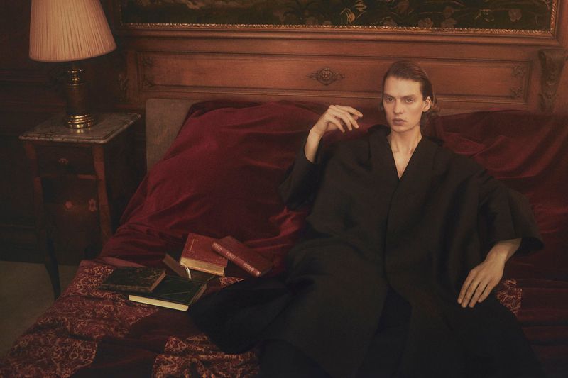 Adult female model in black robe lounging on a red velvet bed with antique books and a bedside table lamp in a warm-toned interior.