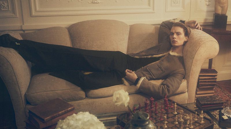 An adult reclining on a sofa in a tailored jacket alongside a chessboard and books in a warm, neutrally lit interior setting.