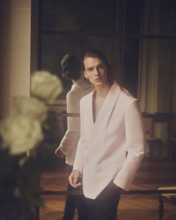 Portrait of a young adult man in a light pink blazer, posing indoors with white roses visible in the foreground.