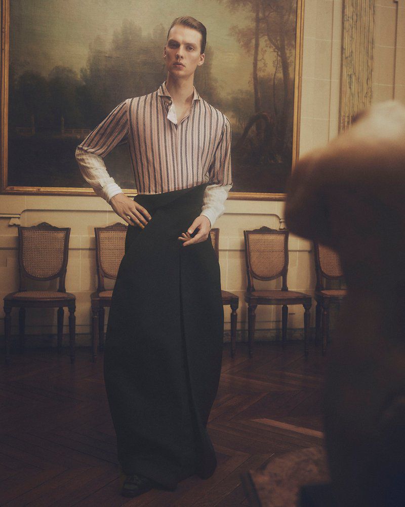 An adult male in a vertically striped shirt and voluminous dark skirt poses indoors in front of a landscape painting in a gilded frame.