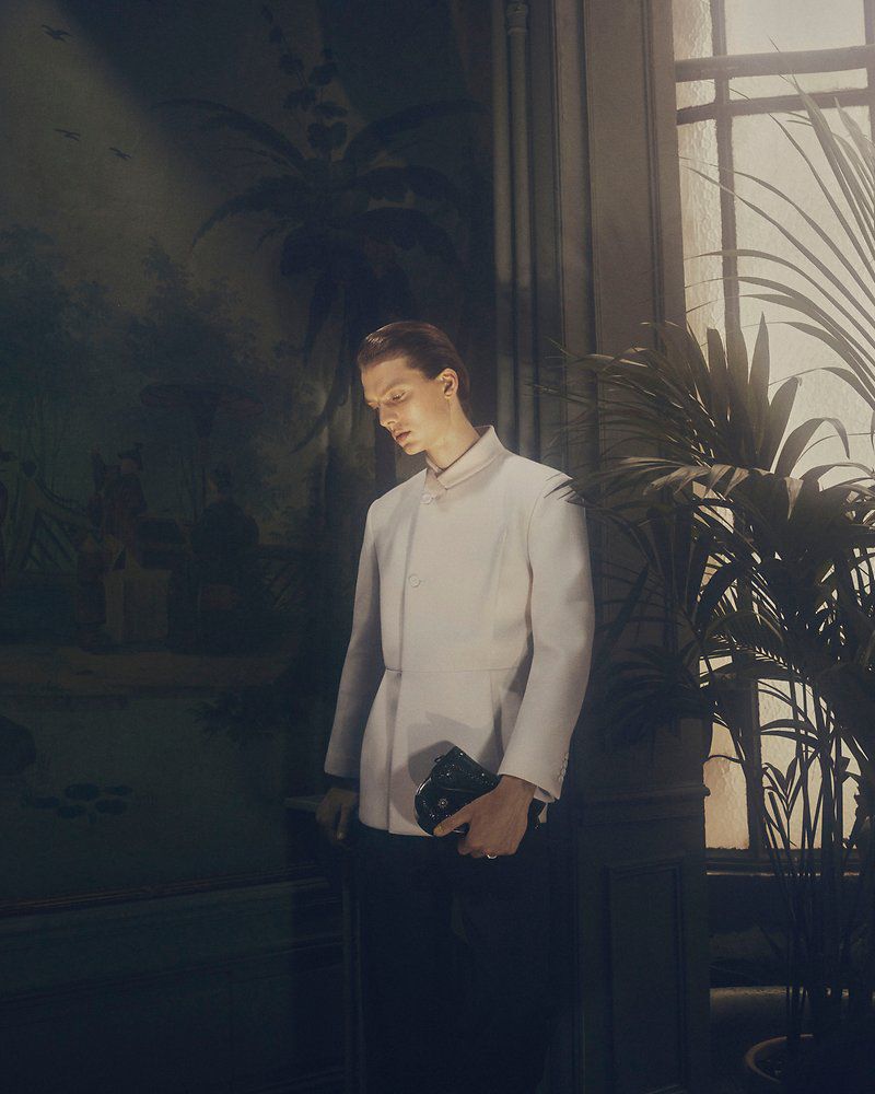Model wearing an off-white tailored jacket holding a black leather clutch while standing near a sunlit window beside a palm plant.