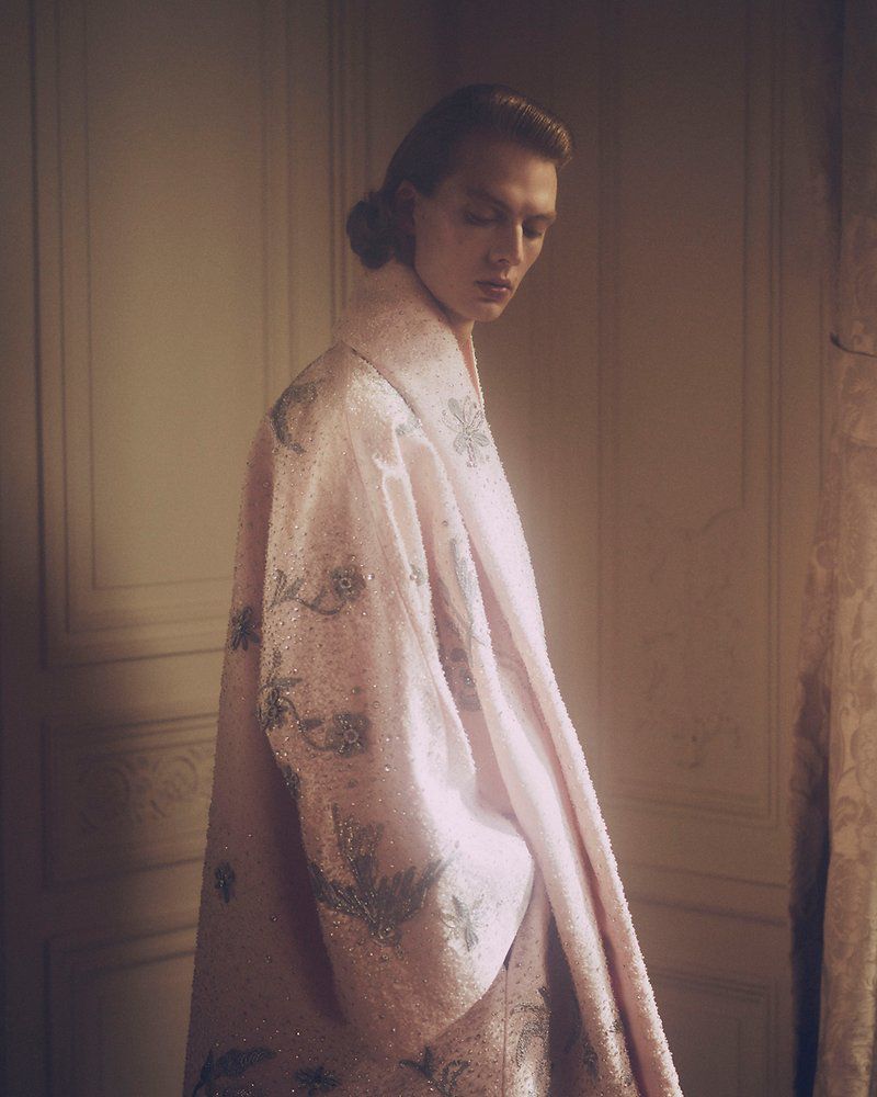 Portrait of an androgynous person wearing a floral pink robe in an indoor setting with neutral wall and door, illuminated by soft lighting.