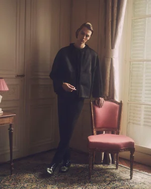 Man standing in an interior leaning on a pink upholstered chair wearing a black jacket and trousers