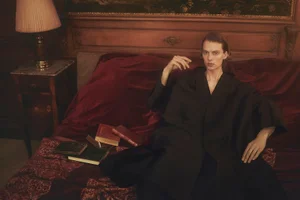 Model in dark coat reclining on a plush red bed with antique books and a bedside lamp in a warm room