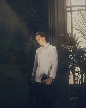 Model wearing an off-white tailored jacket holding a black leather clutch while standing near a sunlit window beside a palm plant.