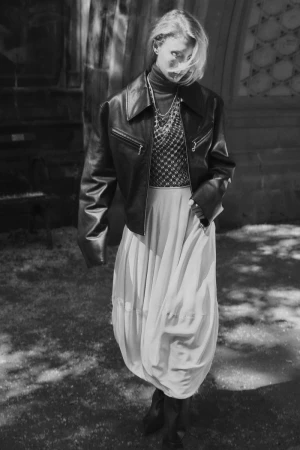 Monochrome portrait of a stylish woman in a leather jacket and full skirt, posing in front of an arched structure, creating a retro fashion moment.