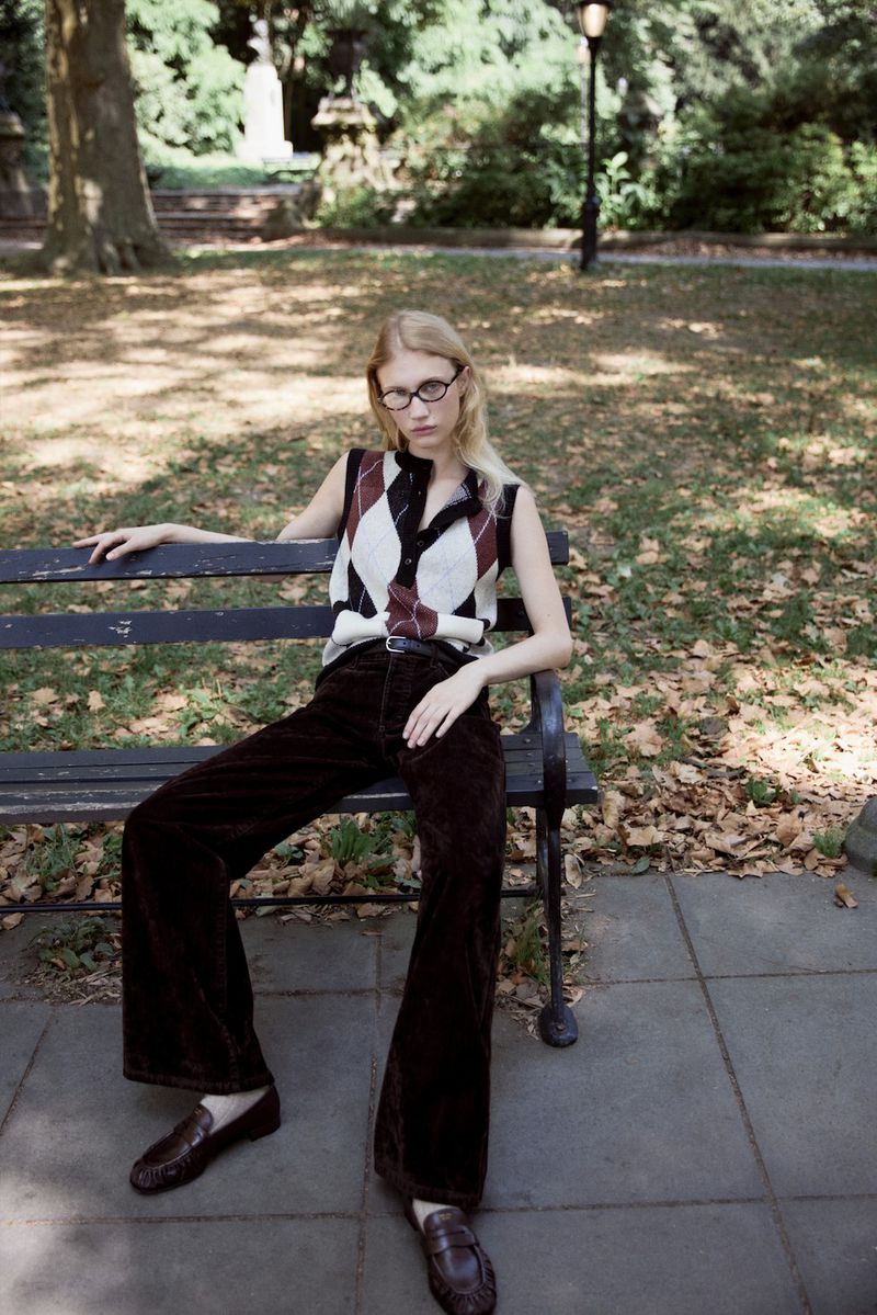 Woman in preppy argyle sweater vest sits on park bench wearing brown corduroy pants and loafers in outdoor setting with trees.