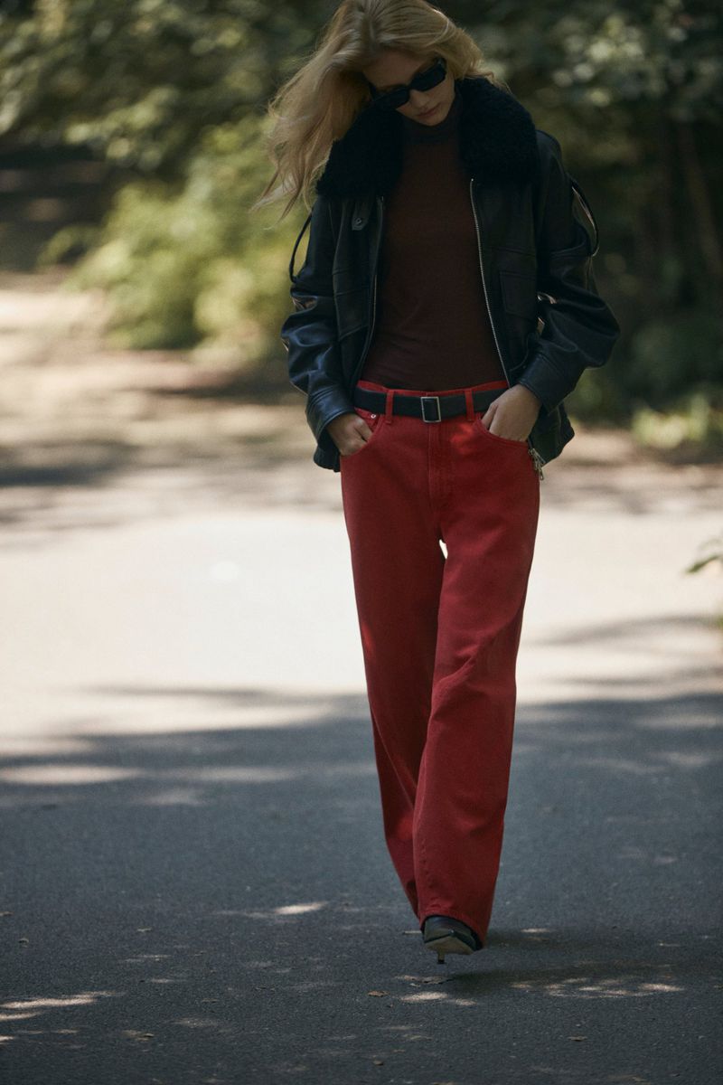 A blonde woman walking in red pants and a black jacket with sunglasses outdoors during the daytime with greenery in the background.