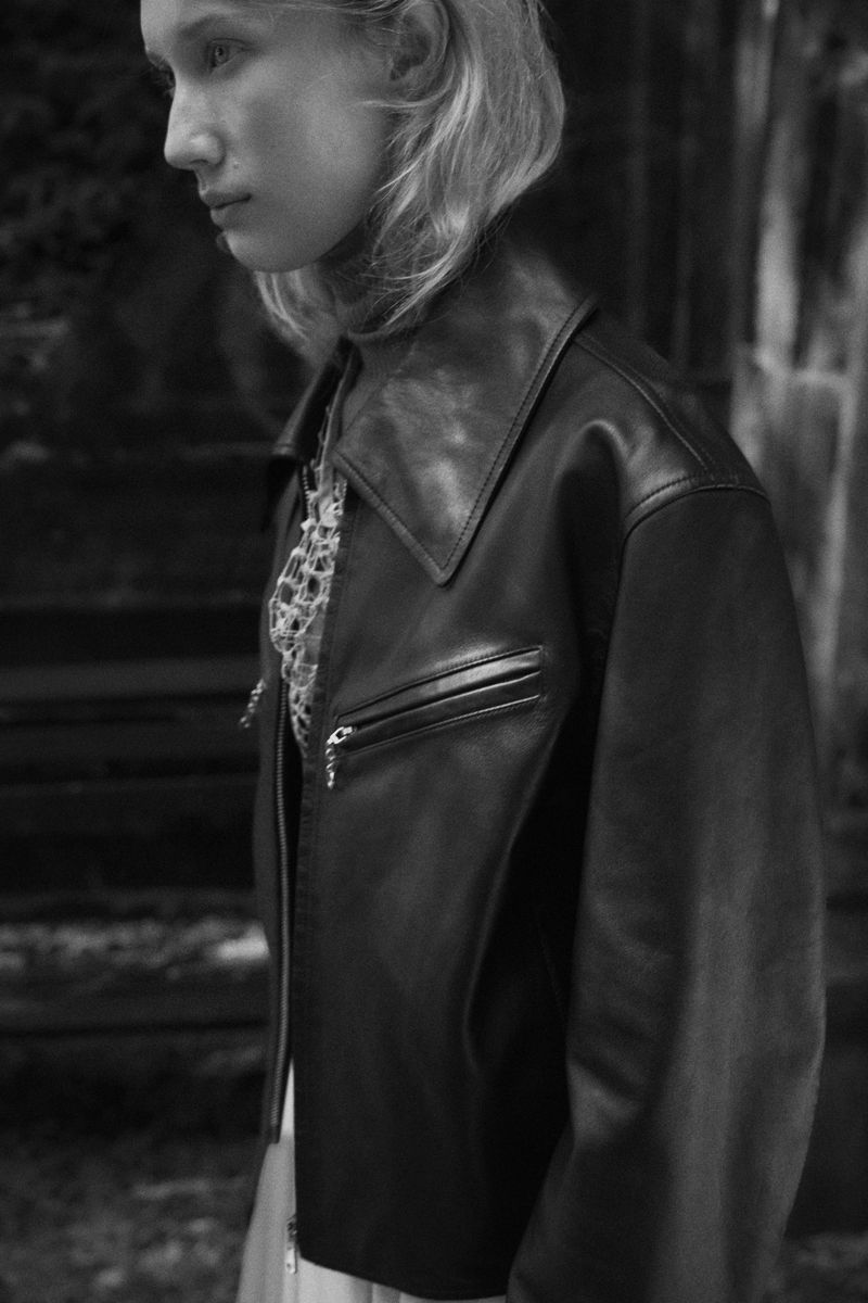 Monochrome close-up of an adult woman in a black leather jacket and patterned top, posing against a dark backdrop.