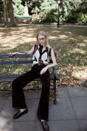 Model seated on a park bench wearing an argyle sweater vest and dark trousers in a park setting.