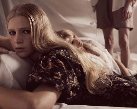 Three fair-skinned adults lie on a white surface, two blonde women in foreground, and one light-skinned person standing in background.