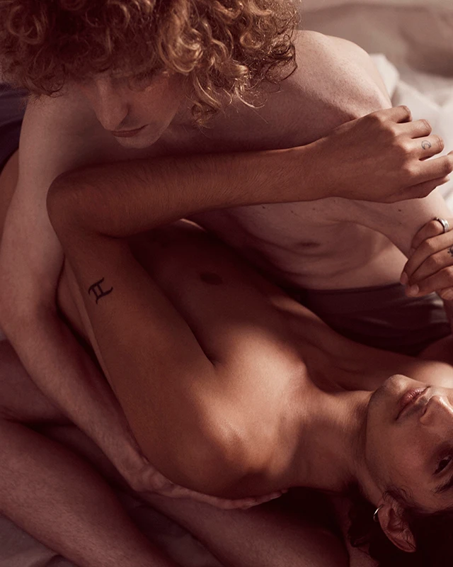Close-up portrait of two shirtless men embracing intimately on a bed with soft indoor lighting by Milos Mlynarik.