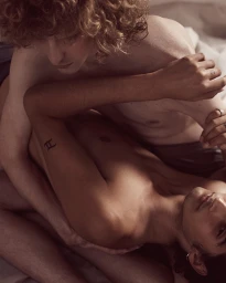 Close-up portrait of two shirtless men embracing intimately on a bed with soft indoor lighting by Milos Mlynarik.