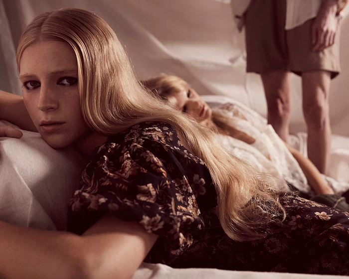 Blonde woman reclining on white bedsheets wearing a dark floral dress with another person and standing figure in the background