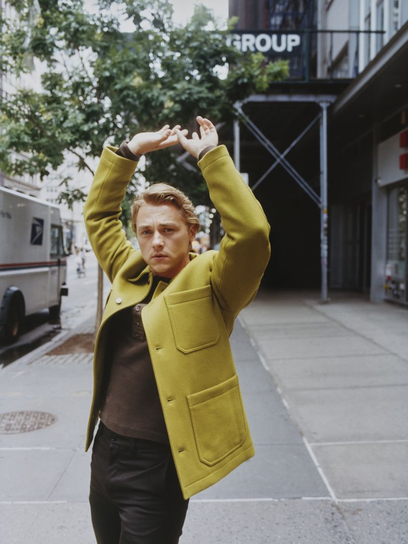 A blond adult man stands in an urban setting wearing a green coat and dark pants, with his arms raised against a background with trees and buildings.