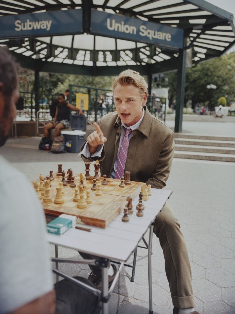 Blond man in suit plays chess in Union Square, New York with an opponent seated across from him in the outdoor scene.