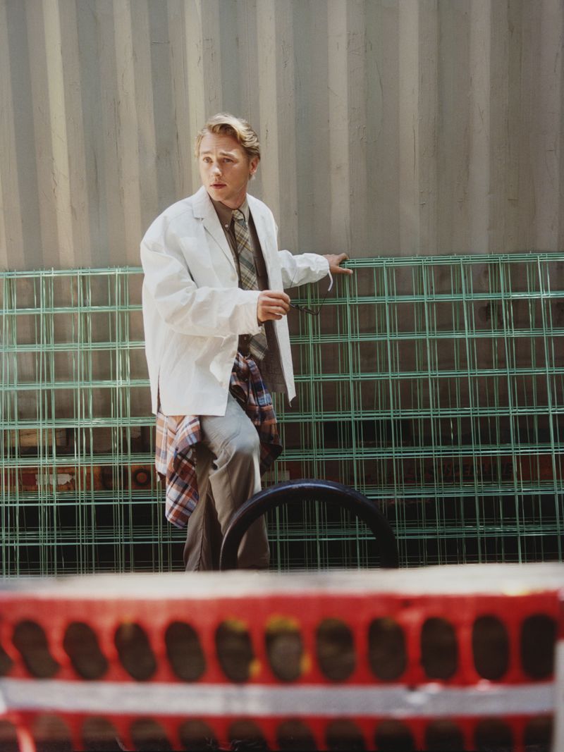 Young man in lab coat standing in front of a green metal grid, wearing a plaid shirt, outdoors with an industrial container backdrop.