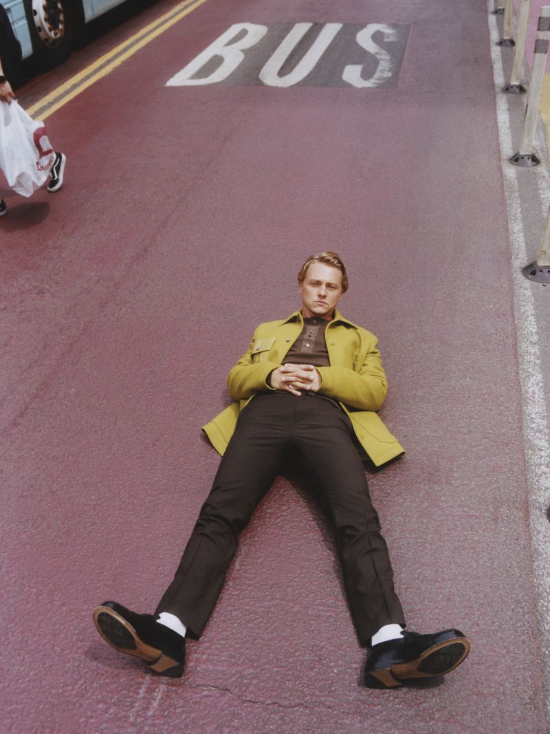Stylish man in yellow coat and black pants lies on a road with 'BUS' marking, creating a striking fashion editorial scene.