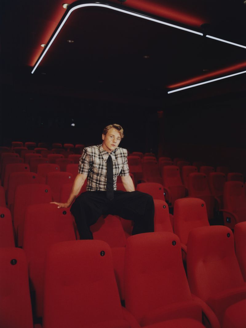 A male model sits in a red-seated movie theatre wearing a plaid shirt, dark pants, and a dark tie in a fashion editorial.