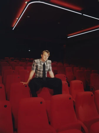 A male model sits in a red-seated movie theatre wearing a plaid shirt, dark pants, and a dark tie in a fashion editorial.