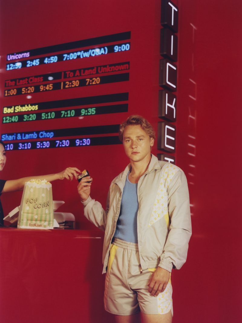 Man buys movie ticket at cinema, holding popcorn. Woman hands him ticket. Red wall with cinema listings and illuminated TICKETS sign.