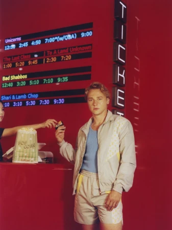 Man buys movie ticket at cinema, holding popcorn. Woman hands him ticket. Red wall with cinema listings and illuminated TICKETS sign.