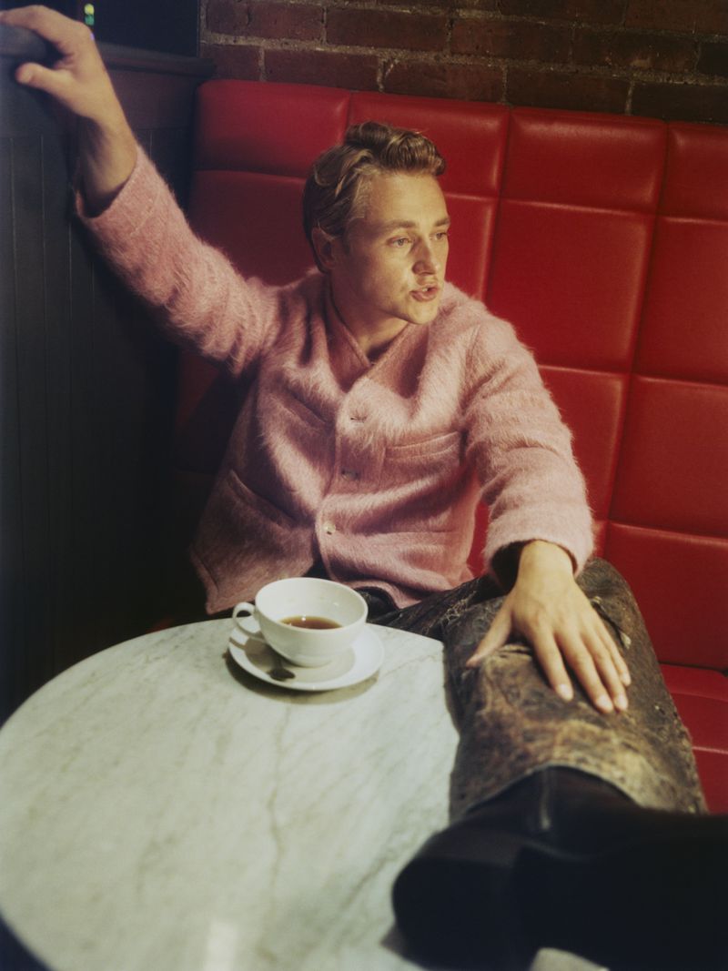 Stylish man in a pink sweater poses at a round table with a cup of coffee, set against a brick wall and red booth seating.