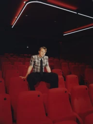 Ben Hardy wearing a plaid short-sleeve shirt and dark trousers seated in a red cinema auditorium for Odda Magazine.