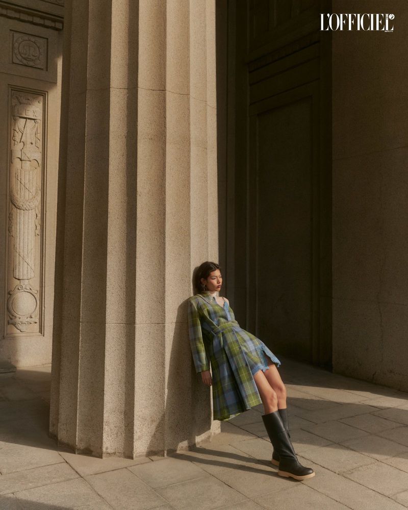 A woman poses in a plaid dress and black boots in front of a column, bathed in sunlight, with the L'Officiel logo visible.